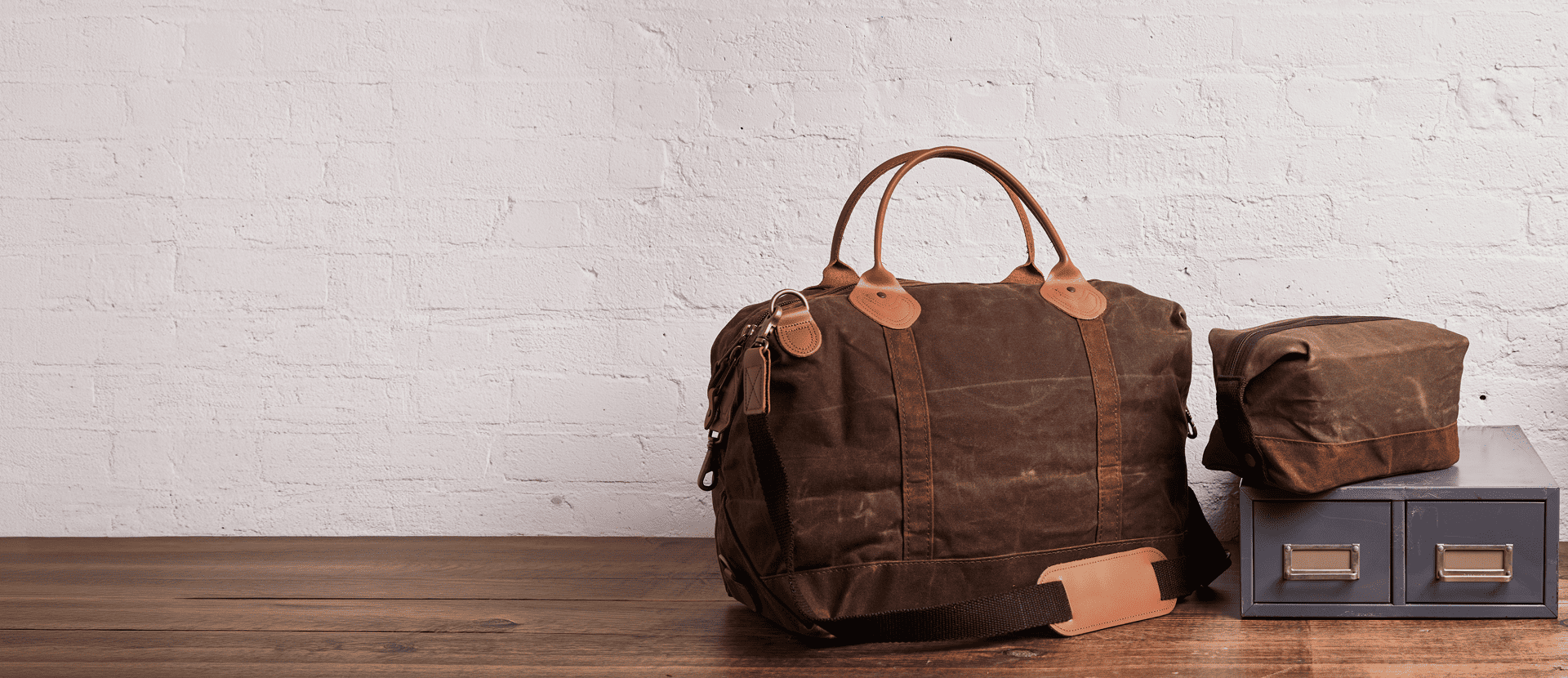 Brown waxed canvas duffel bag and toiletry pouch on a wooden table against a white brick wall