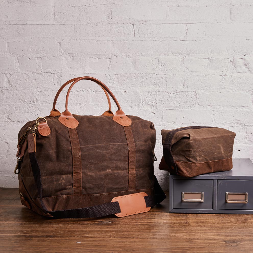 Brown waxed canvas duffel bag and toiletry pouch on a wooden table against a white brick wall