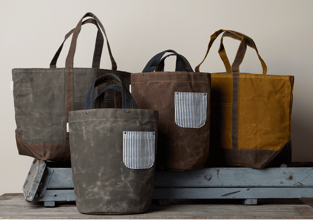 Four waxed canvas tote bags in olive, grey, brown, and mustard on wooden crates